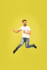 Fototapeta na wymiar Full length portrait of happy jumping man isolated on yellow background. Caucasian male model in casual clothes. Freedom of choices, inspiration, emotions concept. Late for work, has coffee on the run