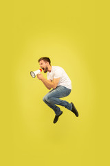 Fototapeta na wymiar Full length portrait of happy jumping man isolated on yellow background. Caucasian male model in casual clothes. Freedom of choices, inspiration, human emotions concept. Calling with mouthpeace.