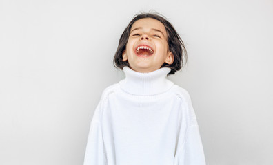 Happy little girl laughing and peeks out of big white knitted sweater, posing over studio light gray studio wall on a holiday. Beautiful child waiting for Christmas presents.