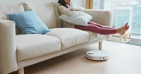 Woman lift up the feet with robotic vacuum on the floor at home - obrazy, fototapety, plakaty