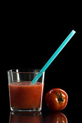 tomato juice in a glass cup on a black background with a stuck blue straw is next to the tomato. On a mirror surface
