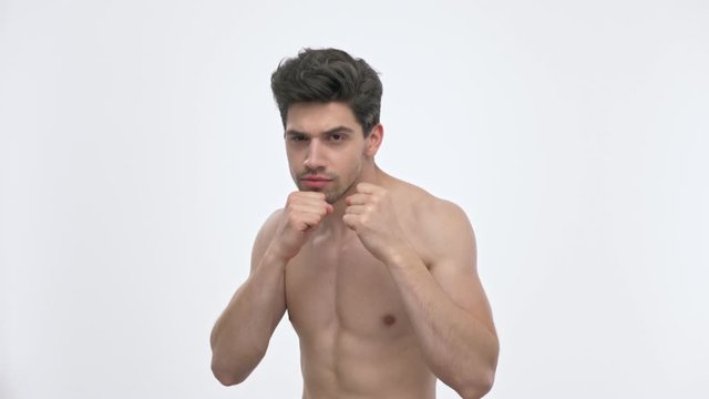 Concentrated young brunette man with naked torso boxing while looking at the camera over white background isolated