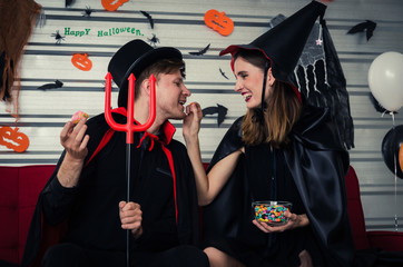 Halloween. couple young caucasian man and woman in vampires and witch clothing feeding sweet snack together and sitting on sofa in halloween party at home, love, holiday and halloween festival concept