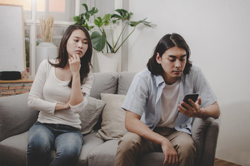 young woman look at her boyfriend from behind who ignore and focused into mobile phone sitting on sofa in living room at home together, upset couple, divorce, family issues and relationship concept