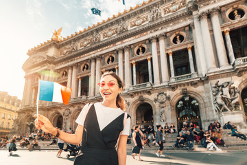 Naklejka premium happy Asian girl traveler with french flag near the main Facade of the Opera Garnier in the historic building of the Academy of music of Paris. Blurred unrecognizable people at the background
