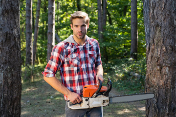 Naklejka na ściany i meble Strong lumberjack with an axe or chainsaw in a plaid shirt. Lumberjack on serious face carries chainsaw. Deforestation is a major cause of land degradation and destabilization of natural ecosystems.