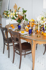 Luxurious elegant wedding decor in autumn style. Wooden vintage table and chairs setting with ceramic plates and silver cutlery. Fresh flowers in a vase, orange pumpkins, deer horns and candles.