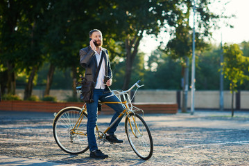 A young stylish bearded businessman in suit going to work by bike. Drinking coffee from a cup to go and talking on a mobile phone on the background of an office building. Takes a working break