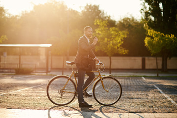 Obraz premium Portrait of handsome young red bearded man in casual clothes using phone, drink coffee and looking away and smiling while leaning on his bike, standing outdoors. Office and business center background