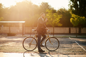 Portrait of handsome young red bearded man in casual clothes using phone, drink coffee and looking away and smiling while leaning on his bike, standing outdoors. Office and business center background