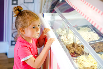 Cute little toddler girl choosing and buying ice cream in a cafe. Happy baby child looking at...
