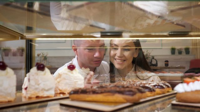 happy young couple chooses tasty fresh cakes standing in glass show case of confectionery shop close view