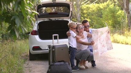 A young active family of travelers in sunglasses view a map near their car on an empty road in the woods. Summer trip by car. Husband, wife and child traveling by car.