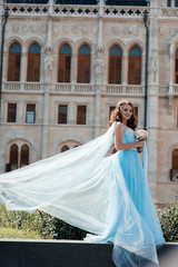 Fototapeta premium Elegant romantic woman in blue sexy dress with flowers standing near the buiding in Prague. Gorgeous girl enjoying. Dressed in blue fluttering dress. Pretty woman with makeup and hairstyle
