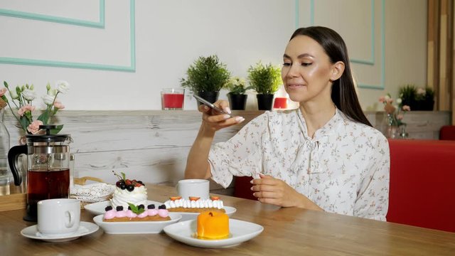 smiling dark haired girl takes picture of tasty natural cakes and tea cups with smartphone at table in cafe