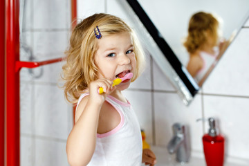 Cute adorable toddler girl holding toothbrush and brushing first teeth in bathroom after sleeping. Gorgeous baby child learning to clean milk tooth. Morning healthy hygiene routine for children