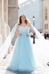 Outdoor lifestyle portrait of pretty young girl on urban background. Pretty smiling joyfully female with fair hair, dressed in blue fluttering dress, looking with satisfaction at camera, being happy.