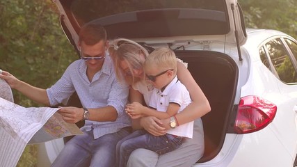 Portrait of a happy family in a car with a map of the area at sunset. The family travels by car. Dad, mom and little son study the route map during a trip in the summer.
