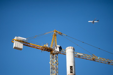 A construction crane erects a tall tower, a civilian passenger plane flies on back of them.