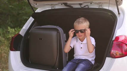 A little cute boy puts on sunglasses sitting in the open trunk of a car before going on vacation. Little cute boy in the trunk of a car with suitcases. Happy family traveling. Summer travel.