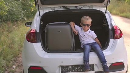 Adorable little kid boy in a sunglasses sitting in car trunk before leaving for summer vacation with his parents. Happy child with suitcases going on journey. Happy family traveling.