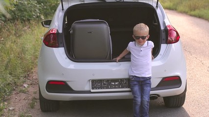 A charming little boy in a white t-shirt, jeans and sunglasses sits in the trunk of a car, in the trunk of a gray suitcase. Family summer trip.