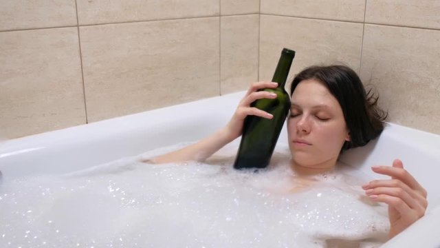 Tired drunk young woman sleeping lying relaxing in bath tub with foam with bottle of wine. Midlife crisis concept. Frustrated tipsy person in depression at home in evening. Disappointment in life.