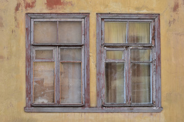 Two small wooden windows in a faded yellow wall covered with old brown spots of paint