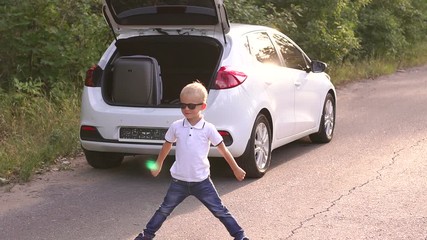 A little funny boy stands on the road near a car in the forest with legs and arms wide apart. Family trip. Happy child with suitcases going on journey. Happy family traveling. Summer travel.