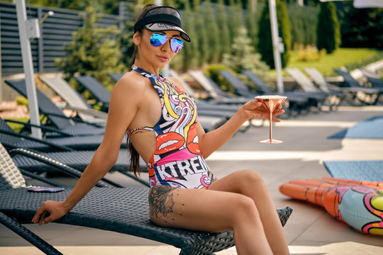 Portrait of a girl having rest and posing sitting on a deck chair near a swimming pool. Dressed in a colorful swimsuit, sun visor and sunglasses.