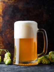 A glass mug of German wheat beer with a large foam cap on a dark background with green hops