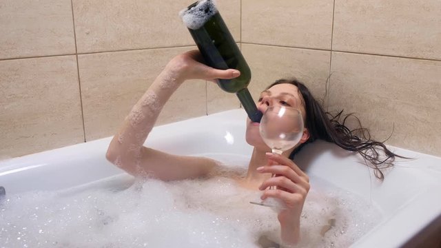 Tired drunk woman lying in foam bath drinking finishes rest of wine from bottle at home in evening. Midlife crisis concept. Frustrated tipsy person in depression. Disappointment life in 30s.