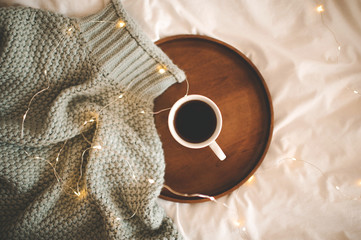 Cup of hot coffee staying on wooden tray with knitted sweater over Christmas lights close up. Top view. Winter holiday season. Good morning.