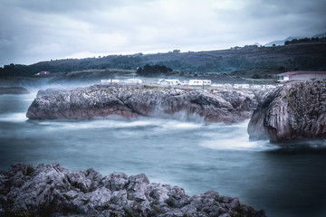 Agua con efecto seda con olas al lado de unos acantilados y monta&ntilde;as de fondo, con un parking para furgonetas camping encima