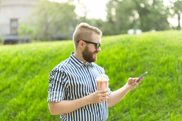 Naklejka na ściany i meble Side view of a stylish young man with glasses and a beard with a milkshake in his hands talking on video with his friends using smartphone and high-speed free Internet in the park on a warm summer day
