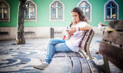 Obraz premium A young woman with a smartphone in her hands sits on a bench in the center of Lisbon