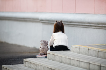 Sad girl with a plush toy sits on the steps of the school stairs with her back to the camera....