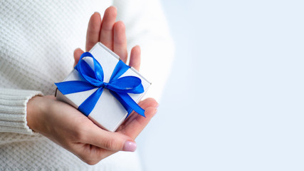Woman in white sweater holding gift box with blue ribbon in hands on white background