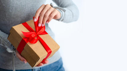 Woman in gray sweater holding gift box with red ribbon in hands on white background