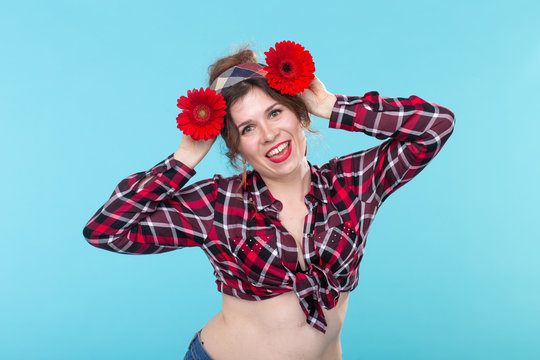 Portrait of a funny smiling pretty young woman in a red checkered shirt substituting red flowers to her head posing against a blue background. Concept of celebration and wonder.