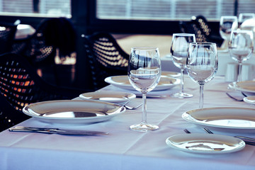 Table setting in restaurant. Empty wine glasses, plates, forks, knifes on the tablecloth. Elegant restaurant interior.