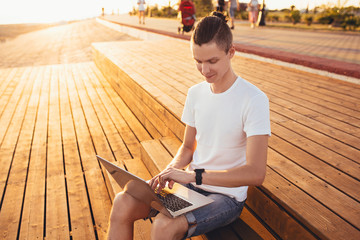 Slim young student boy sits near the beach on wooden benches and admires the view while studying with a laptop on a sunny summer day. Concept of study in the summer