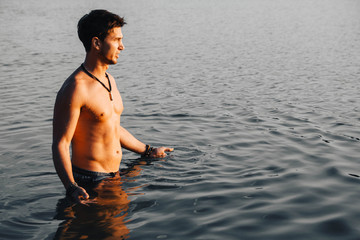 Man with perfect muscular stands in the water at sunset. Place for text or advertising