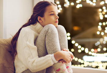 childhood, sadness and people concept - sad beautiful girl in sweater sitting on sill at home...