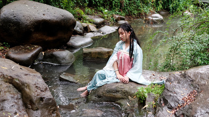 Beautiful Asian woman in cyan costume clothes sitting on big rock near stream with barefoot in water, traditional ancient Chinese beauty, time travel fiction.