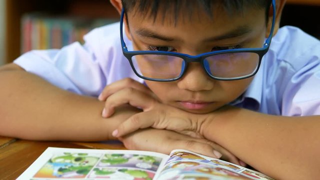 A cute Asian elementary school boy wearing blue glasses in a white school uniform is sitting, enjoying , reading comics in the library.