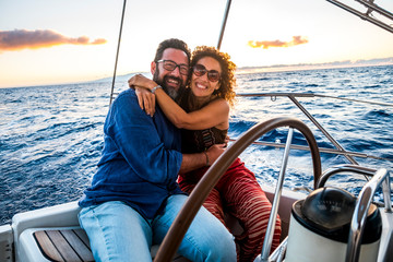 Happy people caucasian adult couple enjoy the sail boat trip on summer holiday vacation - outdoor leisure activity with ocean and sunset in. background - sailing with love and romance - obrazy, fototapety, plakaty