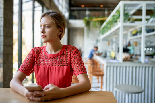 Lifestyle portrait of blonde woman holding her cellphone looking out the window of bright trendy restaurant at large wooden table