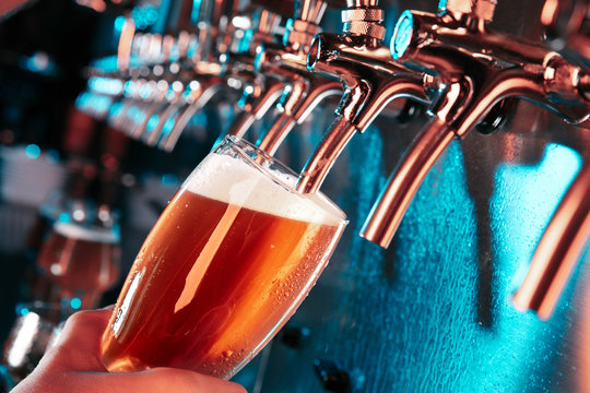 Hand of bartender pouring a large lager beer in tap. Bright and modern neon light, males hands. Pouring beer for client. Side view of young bartender pouring beer while standing at the bar counter.