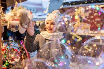 holidays, childhood and people concept - happy little girl choosing christmas balls at market in...